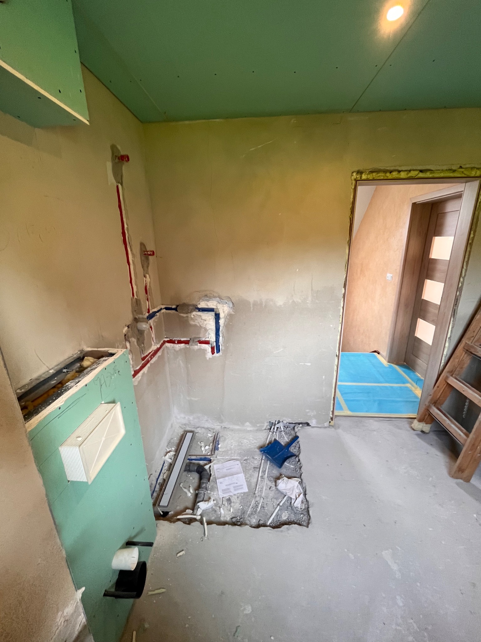 Wnętrze łazienki w trakcie remontu: widoczne rury instalacji wodnej w ścianie, zamontowany stelaż podtynkowy, płyta kartonowo-gipsowa na suficie, zabezpieczona podłoga w sąsiednim pomieszczeniu.