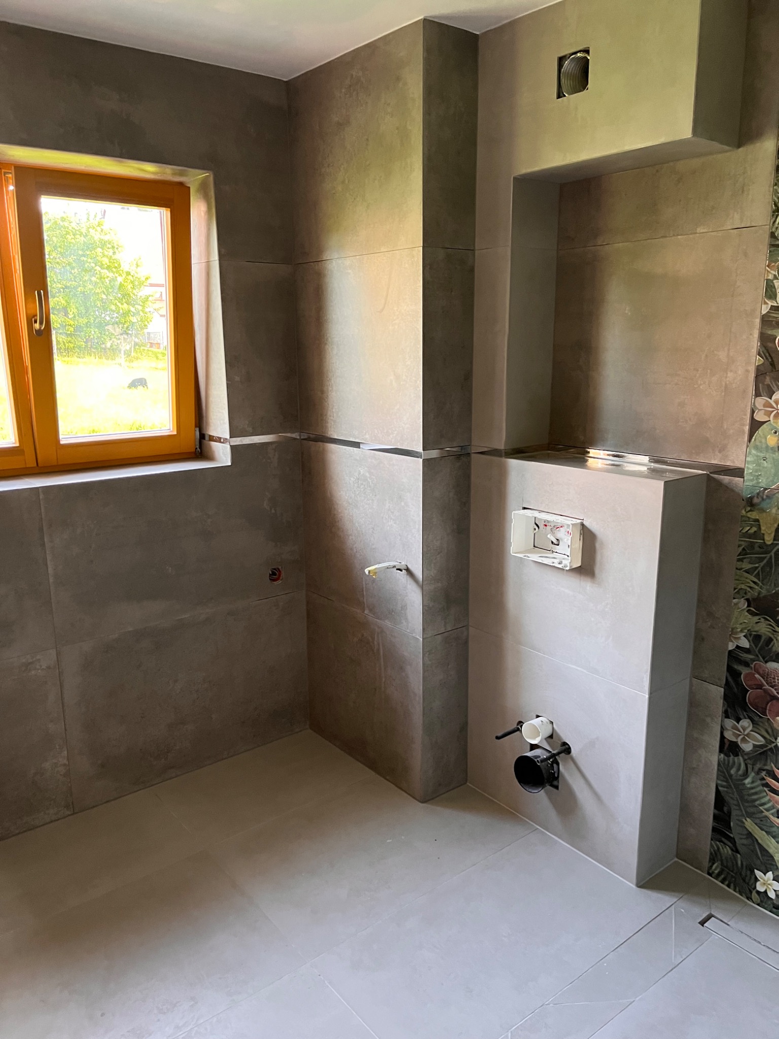 Łazienka w trakcie remontu: szare płytki wielkoformatowe na ścianach i podłodze, drewniane okno z widokiem na zieleń, widoczne elementy instalacji sanitarnej przed montażem, fragment dekoracyjnej...