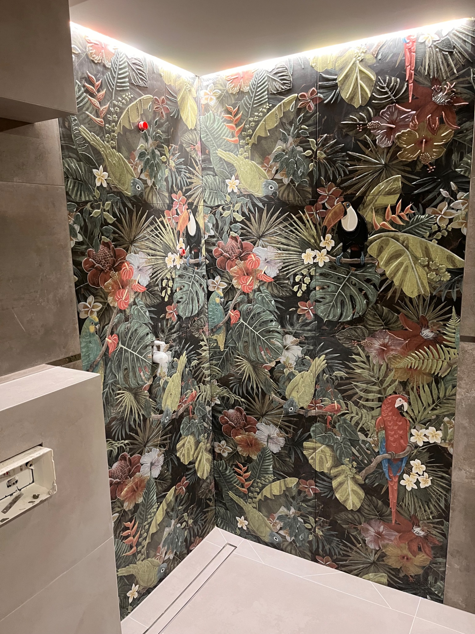 Wykończona ściana prysznicowa z dekoracyjnymi płytkami w tropikalny wzór z motywem papug, tukanów i bujnej roślinności, podświetlona od góry listwą LED.