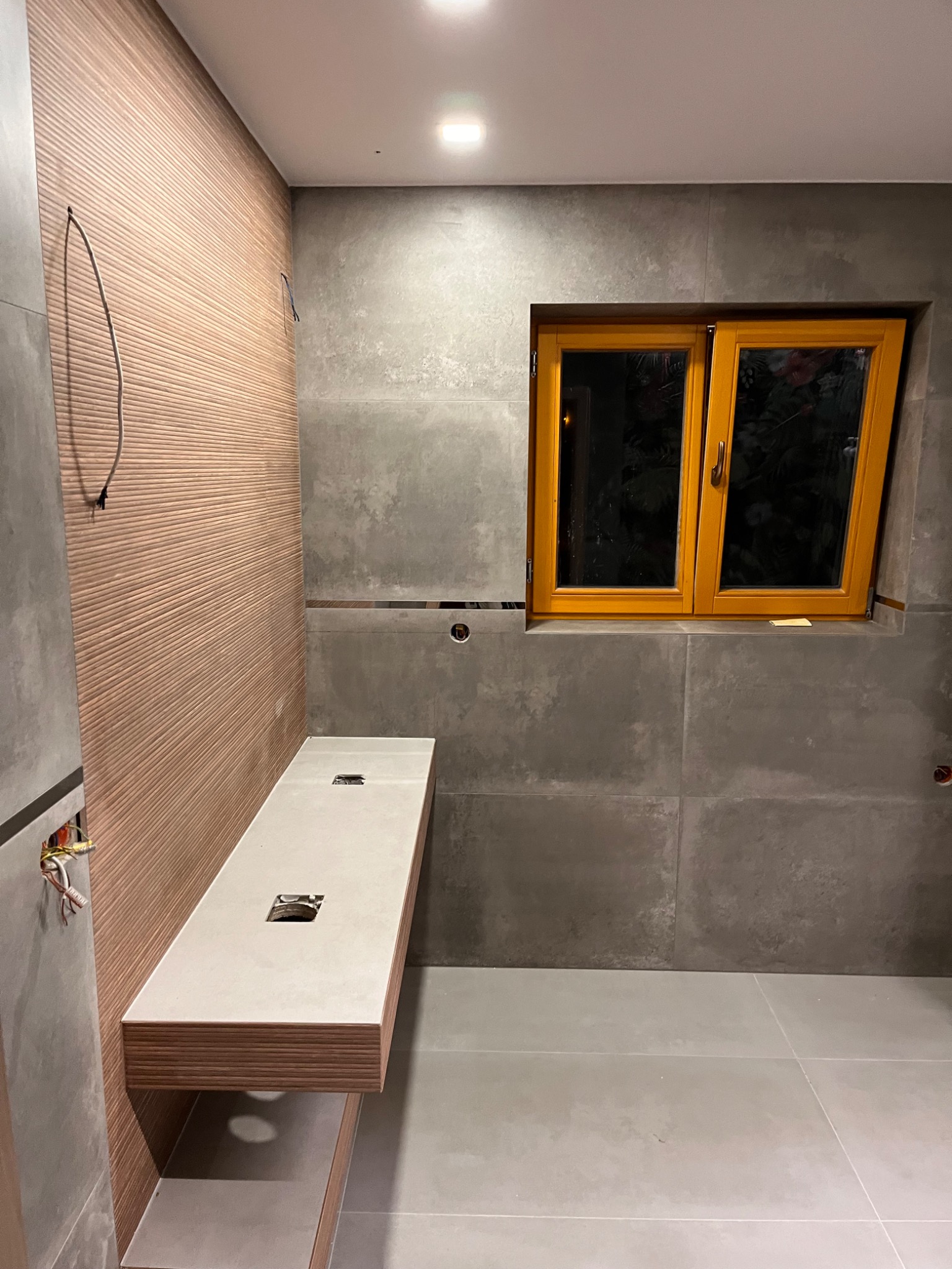 Łazienka w trakcie wykańczania: drewnopodobna ściana z ryflowanym wzorem, szare płytki wielkoformatowe, drewniana rama okienna, podwieszana umywalka z blatem z otworem na odpływ, widoczne kable...