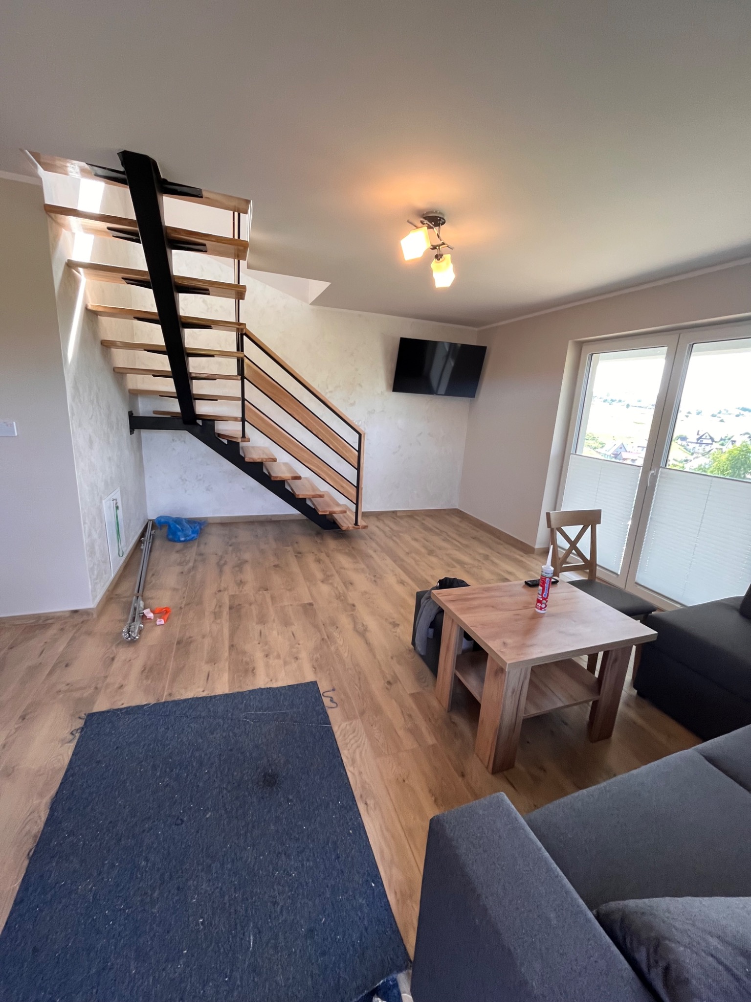 Salon z drewnianą podłogą, schodami z drewnianymi stopniami i czarną konstrukcją, nowoczesnym oświetleniem sufitowym i szarą sofą, częściowo zakrytą ciemną matą ochronną.