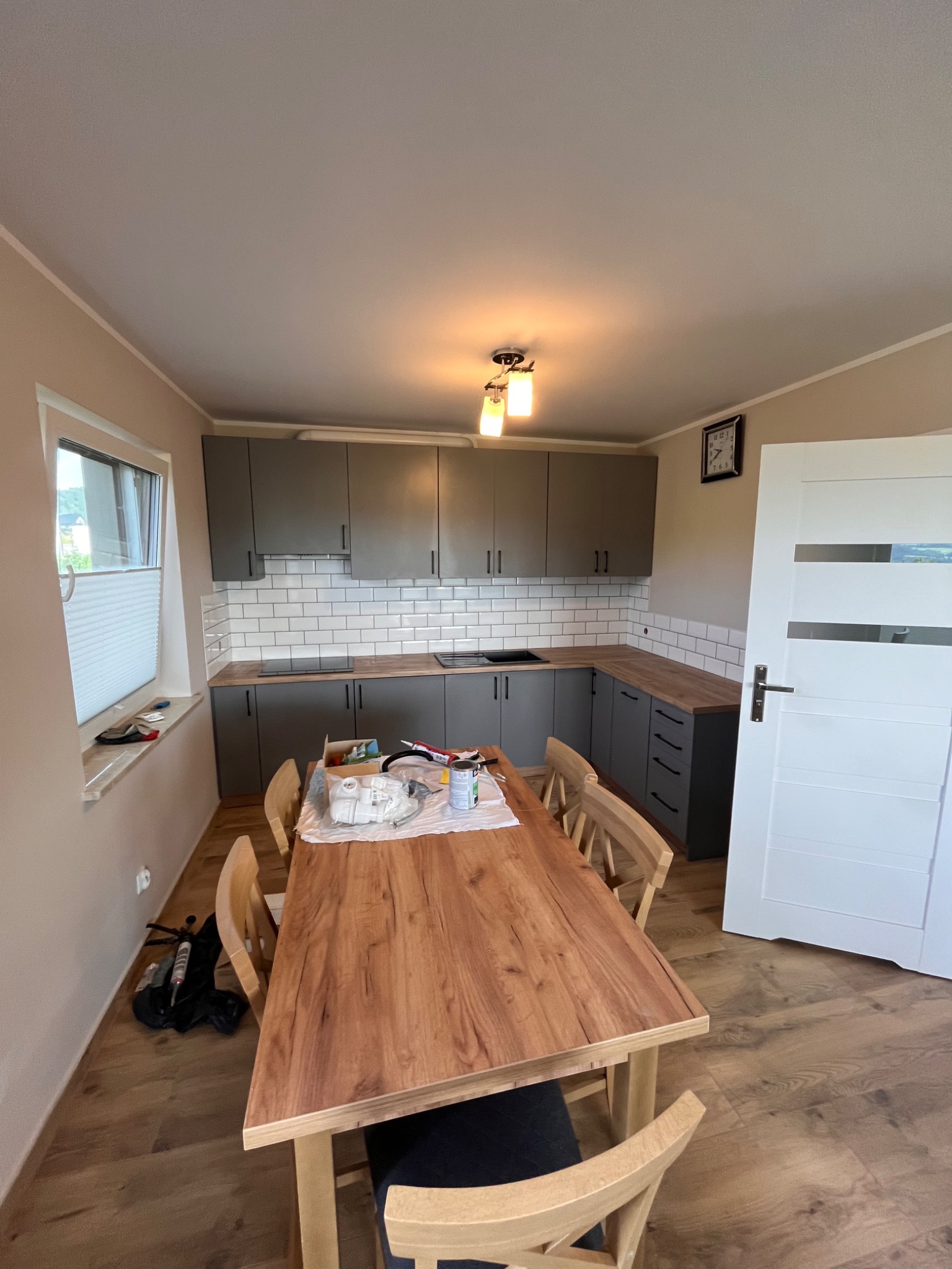 Wnętrze kuchni w trakcie remontu: szare szafki kuchenne z białymi kafelkami na ścianie, drewniany blat, stół z krzesłami, widoczne narzędzia i materiały budowlane na stole i pod oknem.