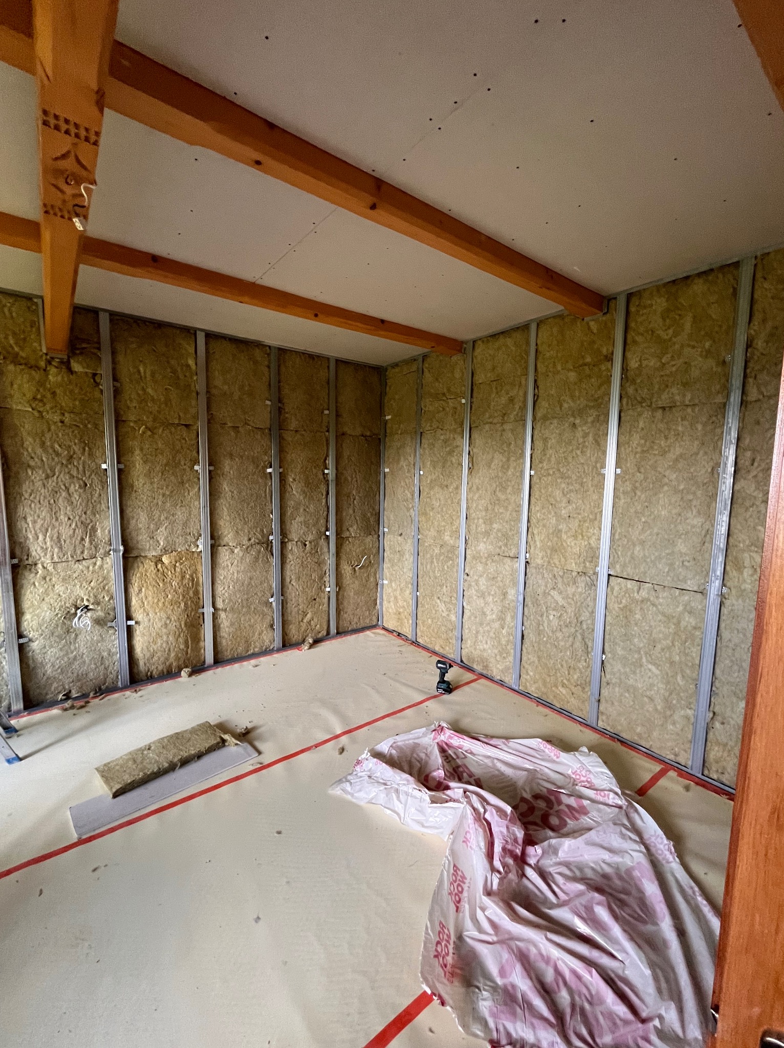 Montaż izolacji z wełny mineralnej w metalowym stelażu, przygotowanie do położenia płyt karton-gips na suficie i ścianach, widoczna wiertarka akumulatorowa i folia ochronna na podłodze.