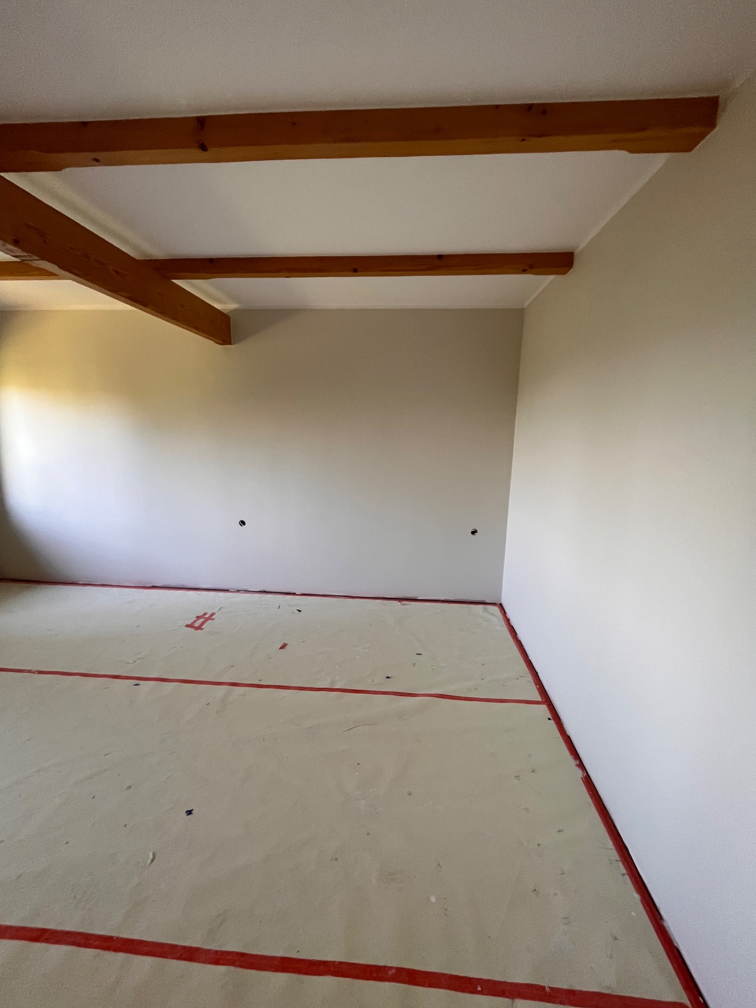 Wnętrze pokoju w trakcie wykończenia, z drewnianymi belkami stropowymi, świeżo pomalowanymi ścianami w odcieniach beżu i zabezpieczoną folią podłogą z czerwoną taśmą.