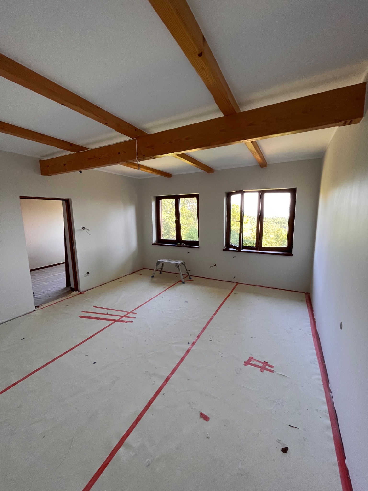 Przygotowania do prac wykończeniowych w pokoju z drewnianymi belkami stropowymi: podłoga zabezpieczona folią i taśmą, widoczne punkty elektryczne, drabina aluminiowa przy oknie.