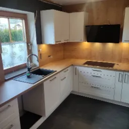 Jasna kuchnia z białymi szafkami i drewnianym blatem, czarny zlew pod oknem, ciemna podłoga, widoczne oświetlenie podszafkowe.