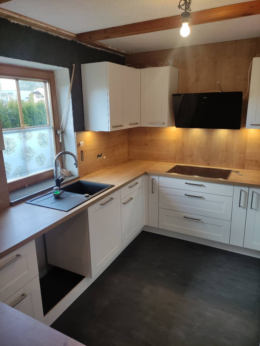 Jasna kuchnia z białymi szafkami i drewnianym blatem, czarny zlew pod oknem, ciemna podłoga, widoczne oświetlenie podszafkowe.