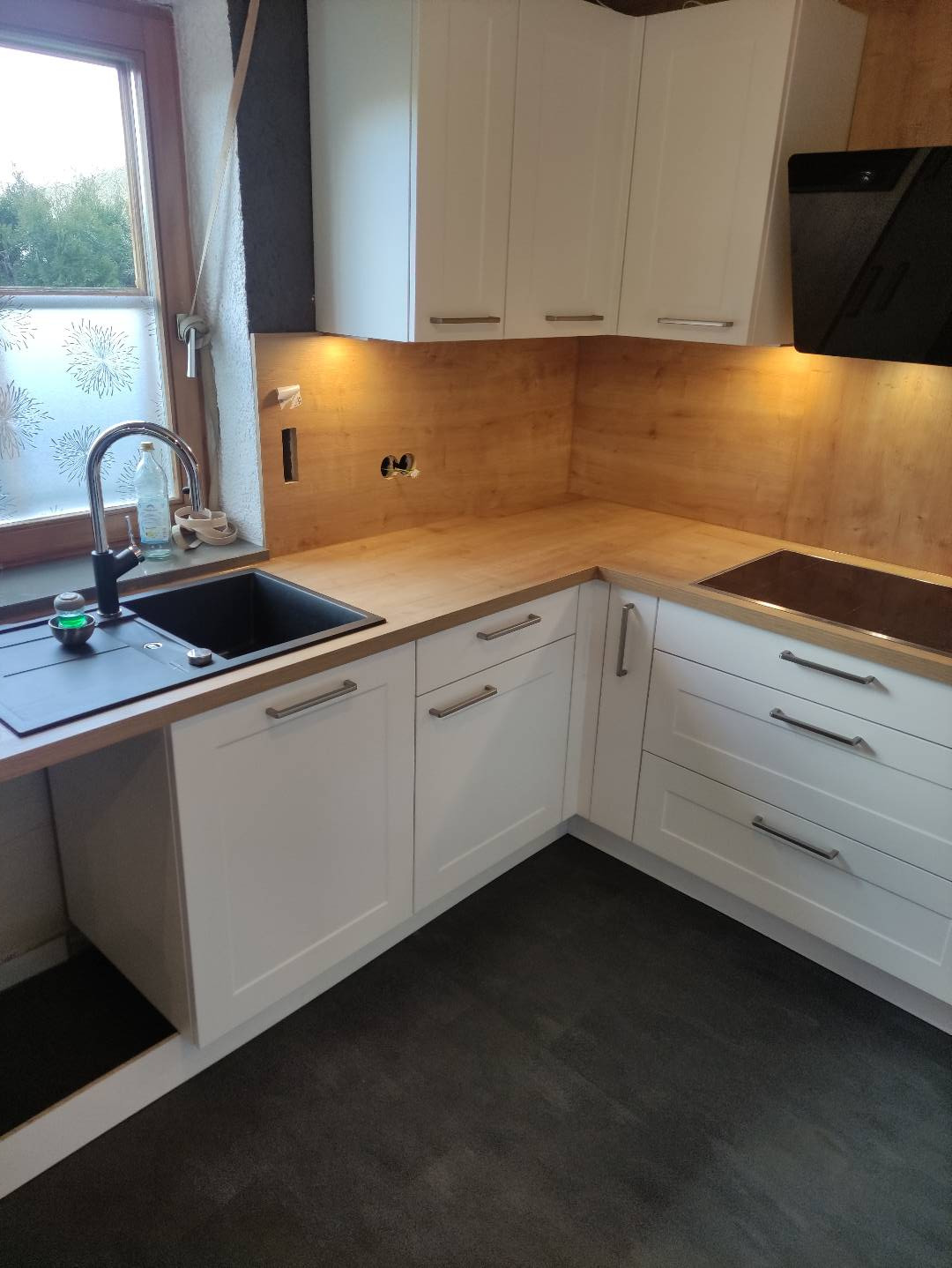 Kąt widzenia na narożną, białą kuchnię z drewnianym blatem, czarnym zlewem i czarną płytą grzewczą, oświetloną od spodu szafek wiszących.