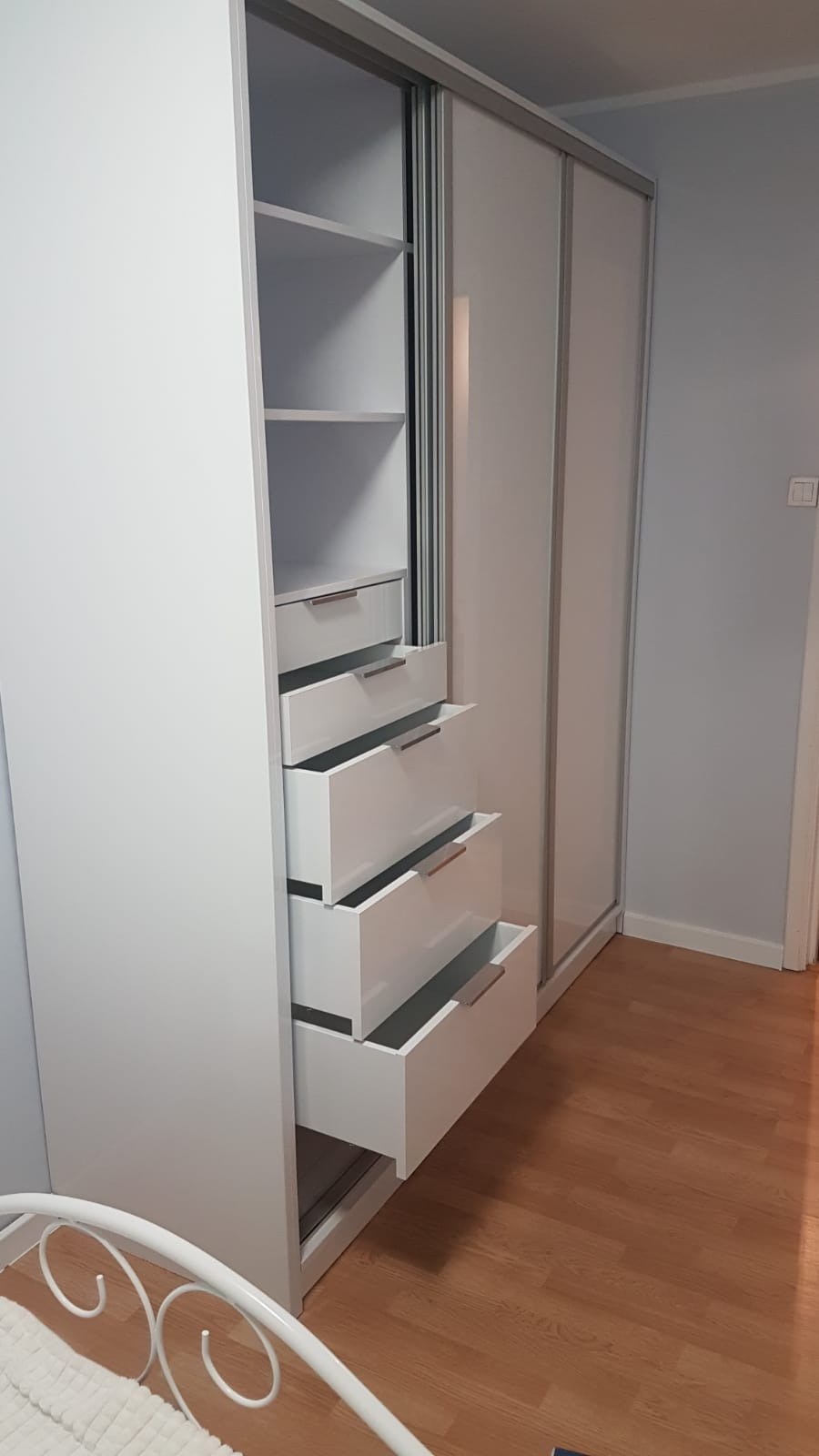 Biała szafa przesuwna z otwartymi szufladami i półkami, widoczne drewniane uchwyty, drewniana podłoga, fragment łóżka w rogu kadru.