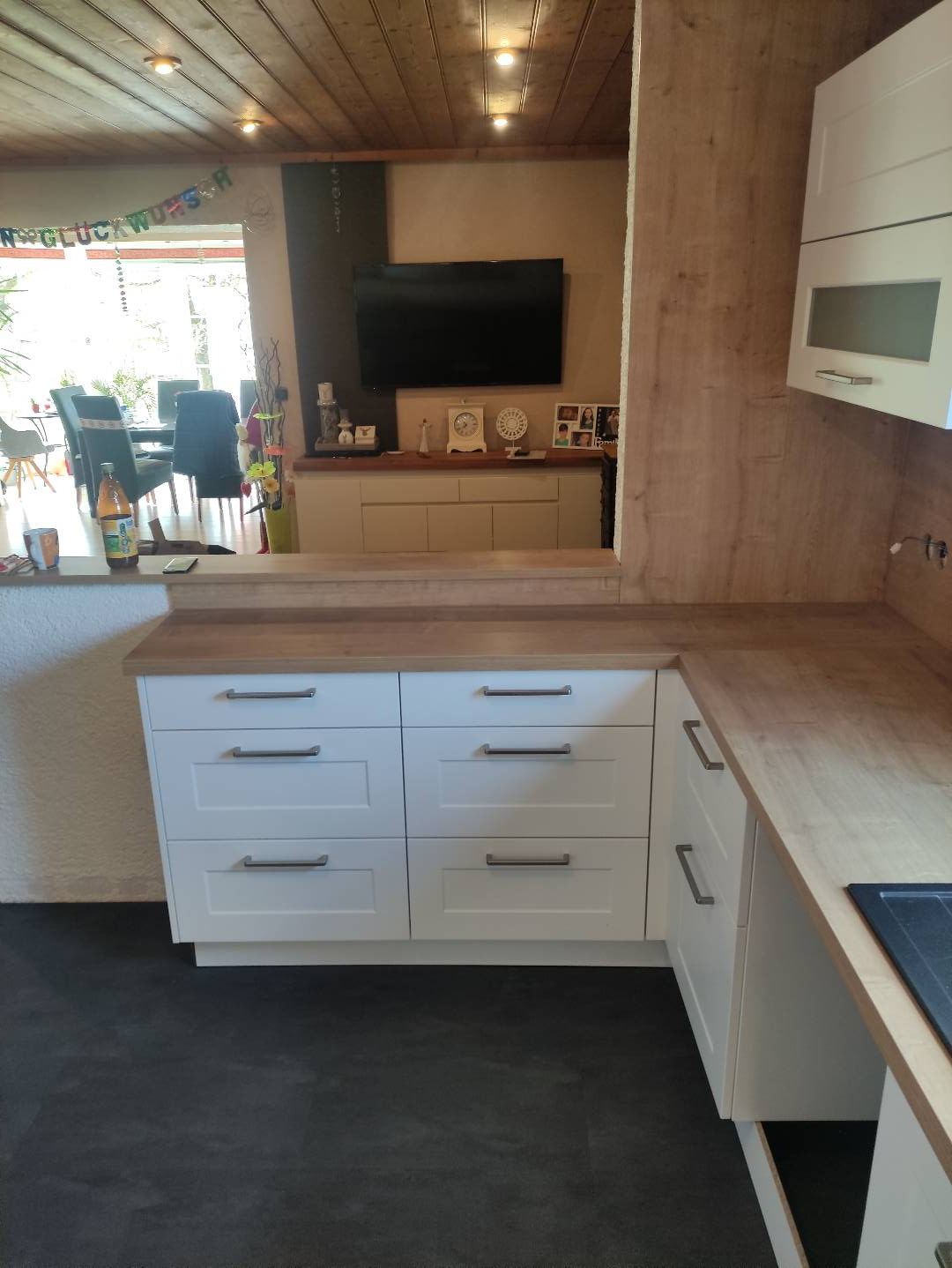 Białe szafki kuchenne z drewnianym blatem, widok na salon z dekoracją urodzinową w tle, czarna podłoga.