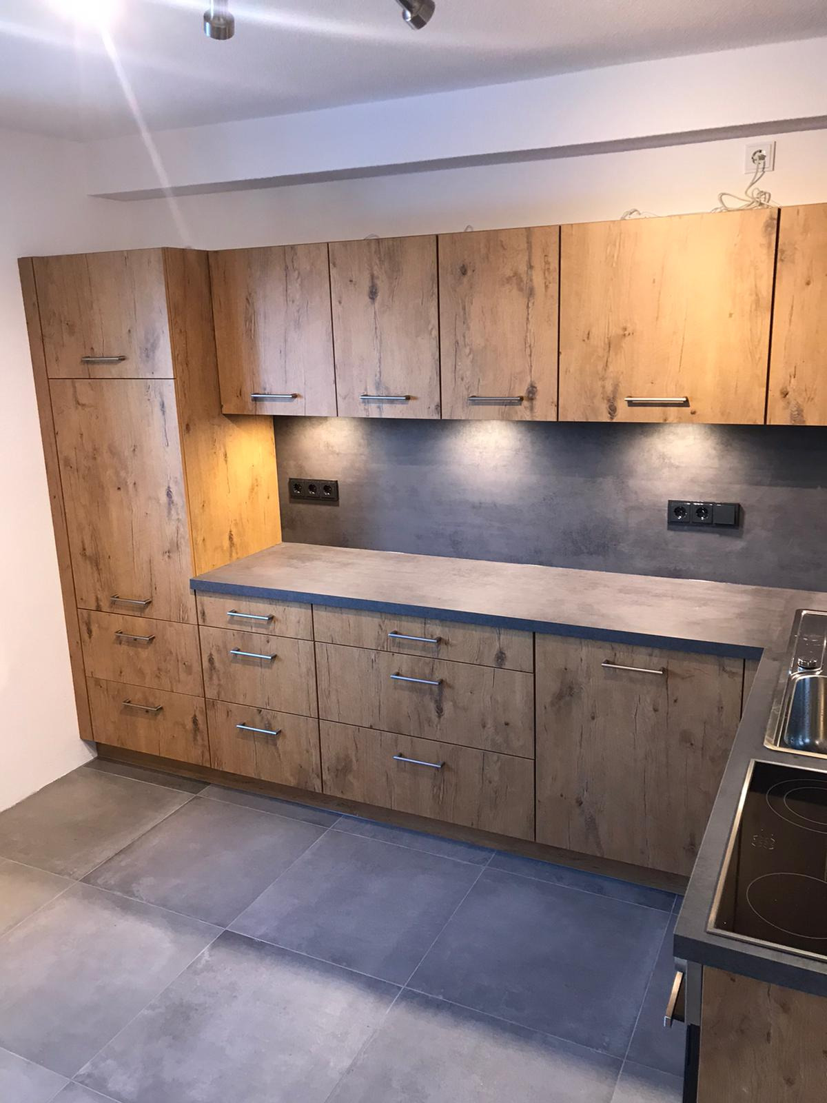 Nowoczesna kuchnia z drewnianymi frontami szafek o wyraźnej strukturze drewna, szarym blatem i szarą ścianą nad blatem, oświetlona punktowymi lampkami podszafkowymi.