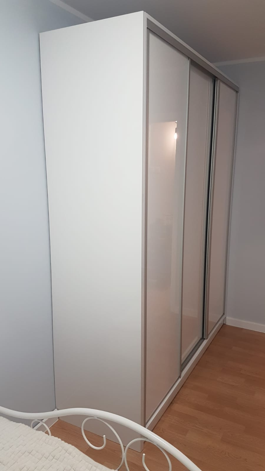 Biała szafa wnękowa z przesuwnymi drzwiami i srebrnymi ramami, widoczny fragment metalowego łóżka w rogu, na podłodze panele drewniane