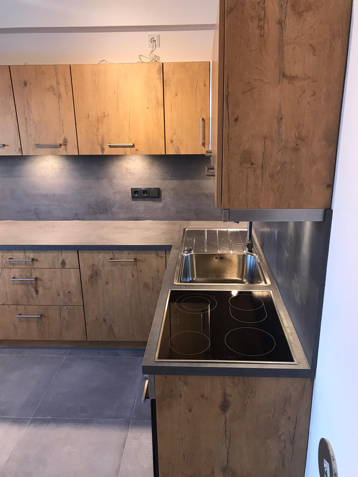 Nowoczesna kuchnia z drewnopodobnymi frontami, szarym blatem i płytą indukcyjną, oświetlenie punktowe pod szafkami wiszącymi, widok na zlew i fragment ściany.