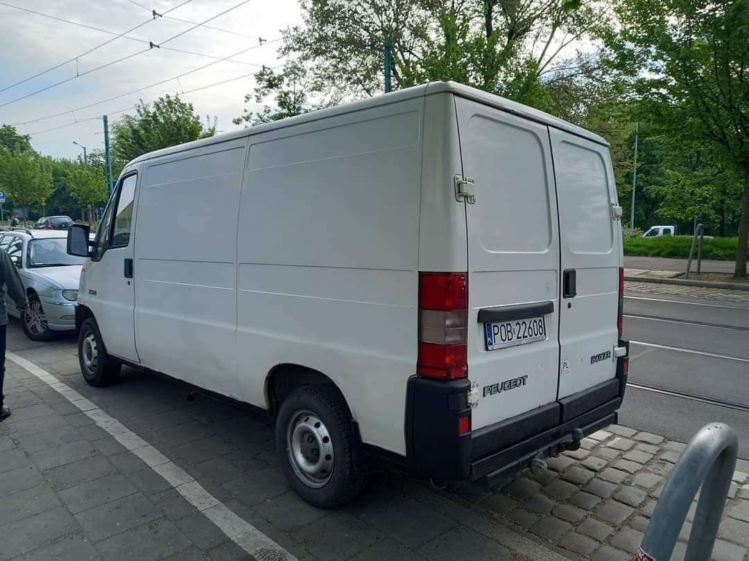 Biały samochód dostawczy Peugeot Boxer zaparkowany na chodniku, widok z tyłu, z widoczną tablicą rejestracyjną i zarysami drzew w tle.