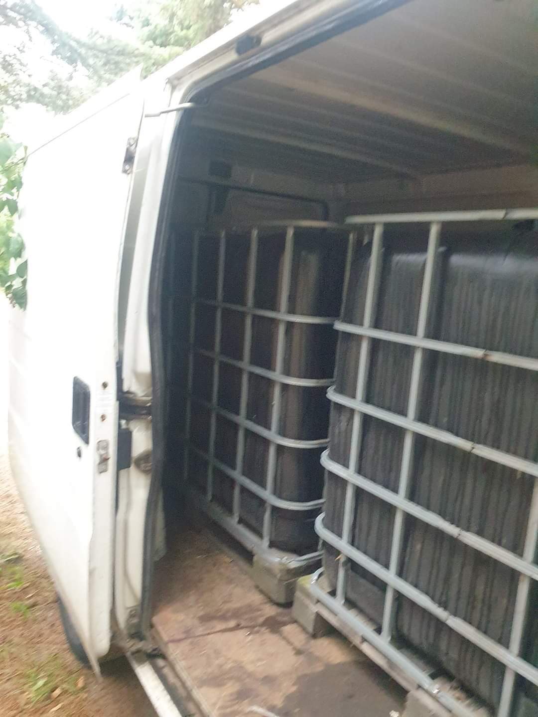 Wnętrze białego busa z otwartymi drzwiami bocznymi, załadowane dwoma dużymi czarnymi pojemnikami IBC na paletach, widok z zewnątrz na częściowo zacieniony teren.