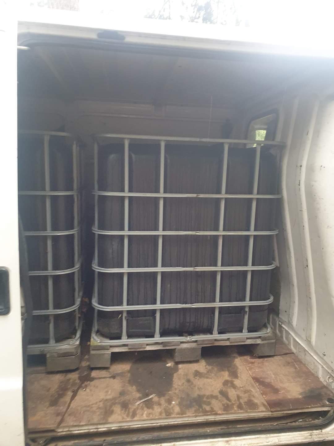 Dwa czarne kontenery IBC na paletach umieszczone w przestrzeni ładunkowej białego busa, widok z tyłu, słabe oświetlenie.