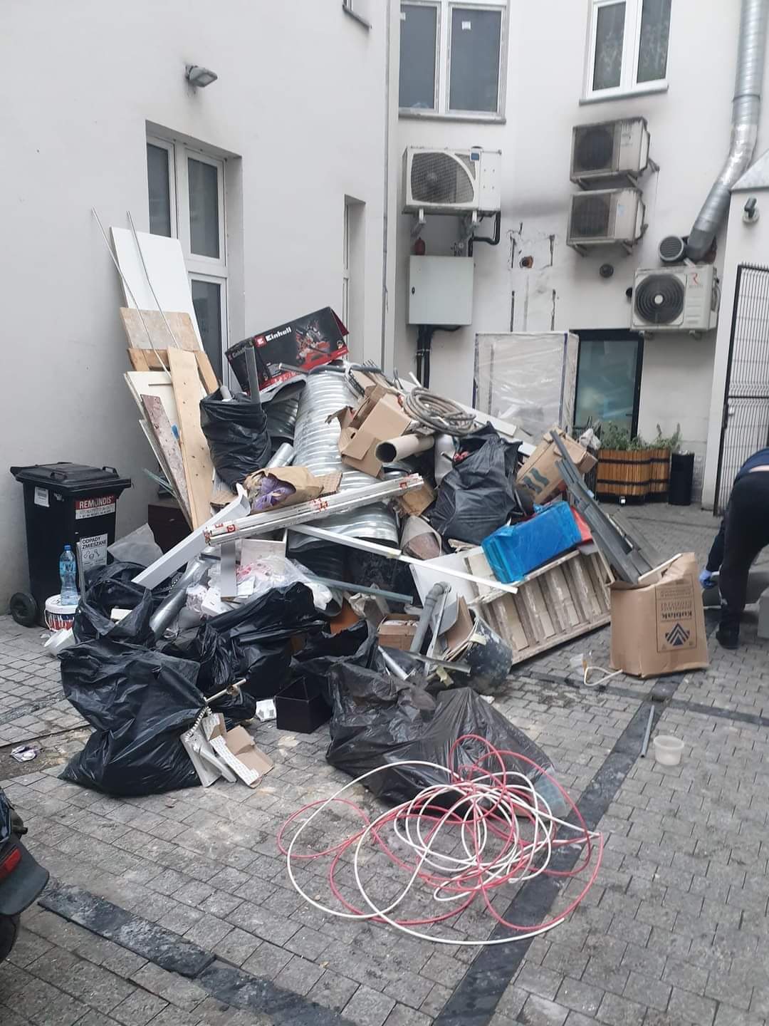 Sterta gruzu budowlanego i odpadów w czarnych workach oraz kartonach na brukowanym dziedzińcu, z widocznymi elementami rozbiórki, takimi jak fragmenty płyt, rury i przewody, na tle ściany budynku...