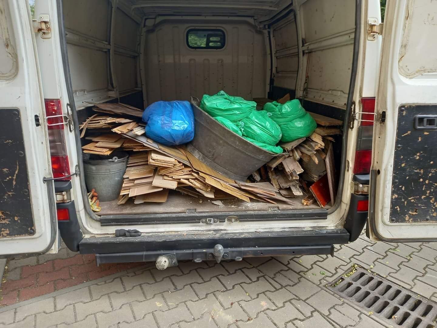 Wnętrze brudnego, białego busa z otwartymi drzwiami załadowane gruzem, drewnem, metalową balią i workami na śmieci w kolorze zielonym i niebieskim.