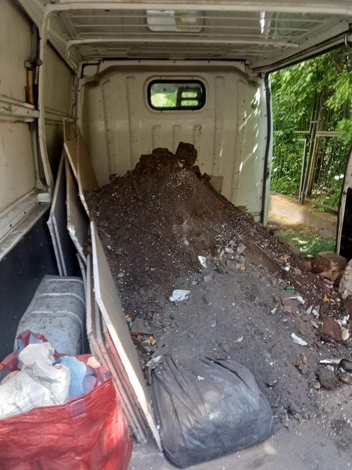 Wnętrze białego busa wypełnione stertą ziemi, gruzu i odpadów budowlanych, widoczne płyty gipsowe, worki na śmieci i torba z materiałem.