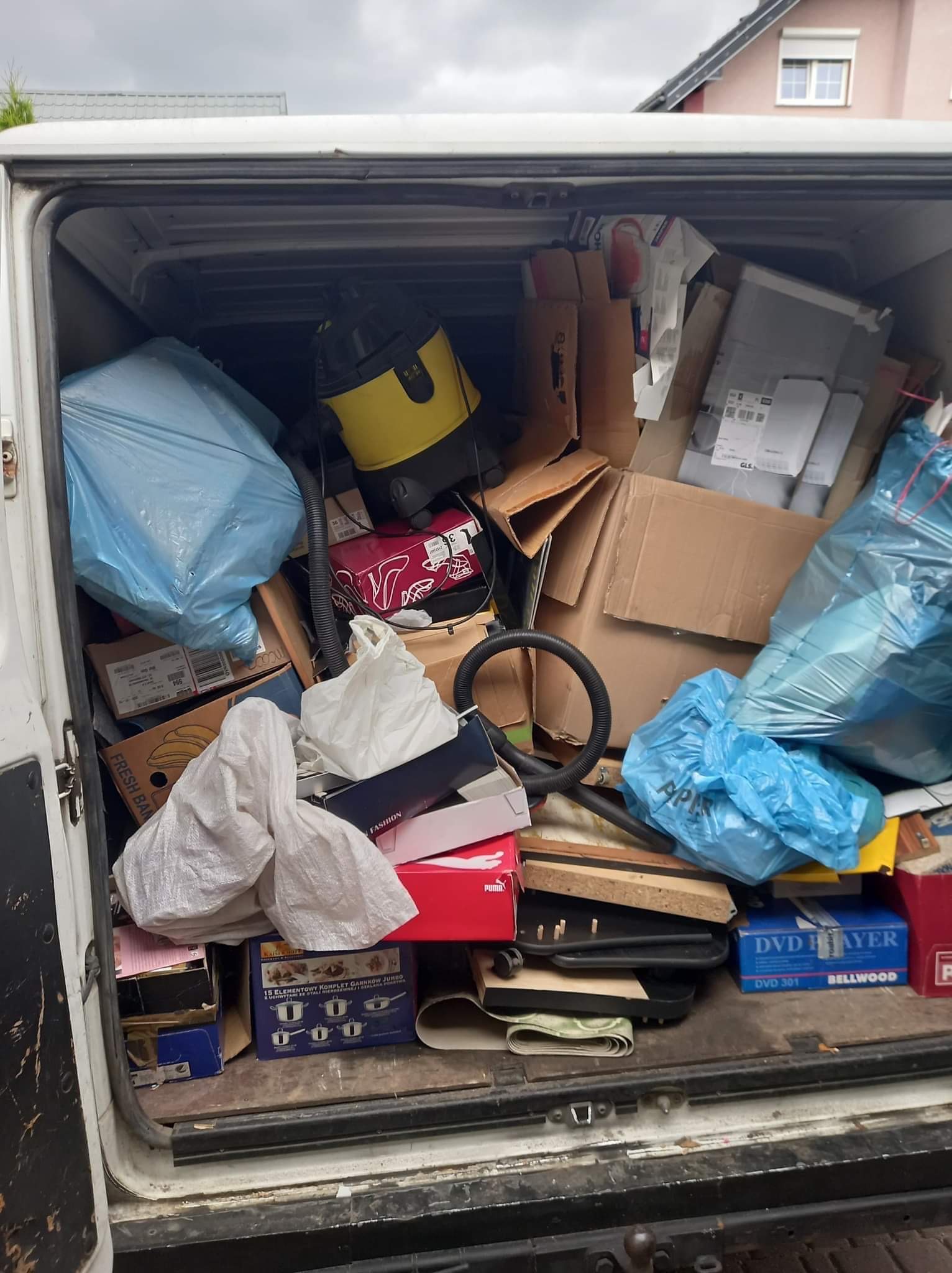 Wnętrze furgonetki wypełnione różnorodnymi przedmiotami, w tym worki na śmieci, kartony, odkurzacz przemysłowy oraz pudełka po butach i garnkach. Widoczny bałagan sugeruje transport różnego rodzaju...