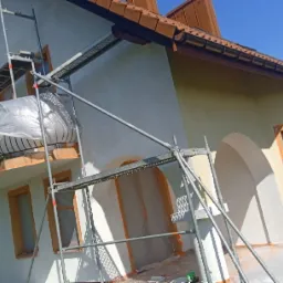 Dom jednorodzinny w trakcie prac elewacyjnych, osłonięte okna i balkon folią malarską, rusztowanie na pierwszym planie, świeżo nałożony tynk na jednej ze ścian.