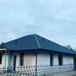 Dom jednorodzinny w trakcie budowy, z ciemną dachówką na nowym dachu, widoczne niezabudowane otwory okienne i ogrodzenie posesji.