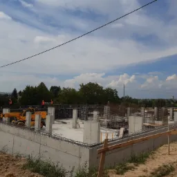 Wykop fundamentów domu z betonowymi ścianami i zbrojeniem, widoczny żółty podnośnik teleskopowy i pracownicy w pomarańczowych kamizelkach na tle błękitnego nieba z chmurami.