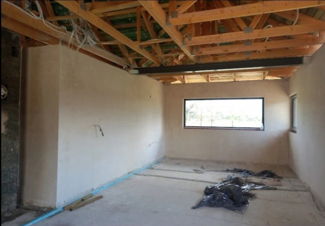 Wnętrze w trakcie prac wykończeniowych: surowe ściany pokryte tynkiem, widoczna drewniana konstrukcja dachu, niebieskie rurki przy podłodze, okno z widokiem na zieleń.