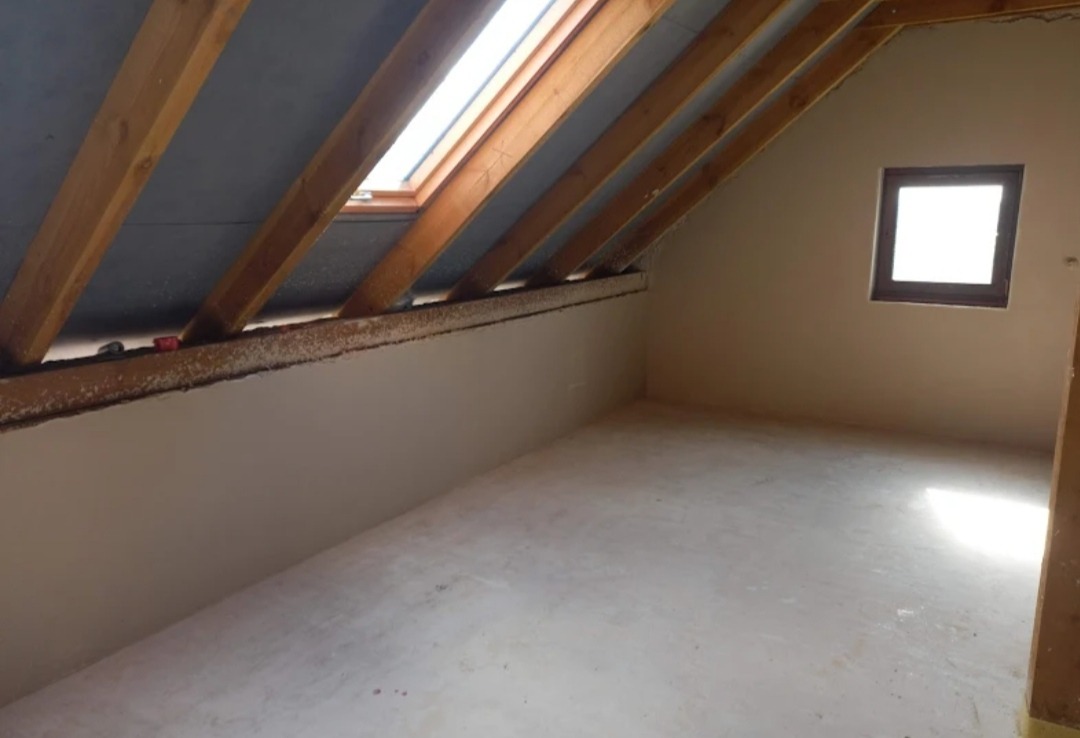 Poddasze po tynkowaniu maszynowym, widoczne drewniane belki stropowe, okno dachowe i okno w ścianie szczytowej; ściany i podłoga pokryte świeżym, jasnym tynkiem.