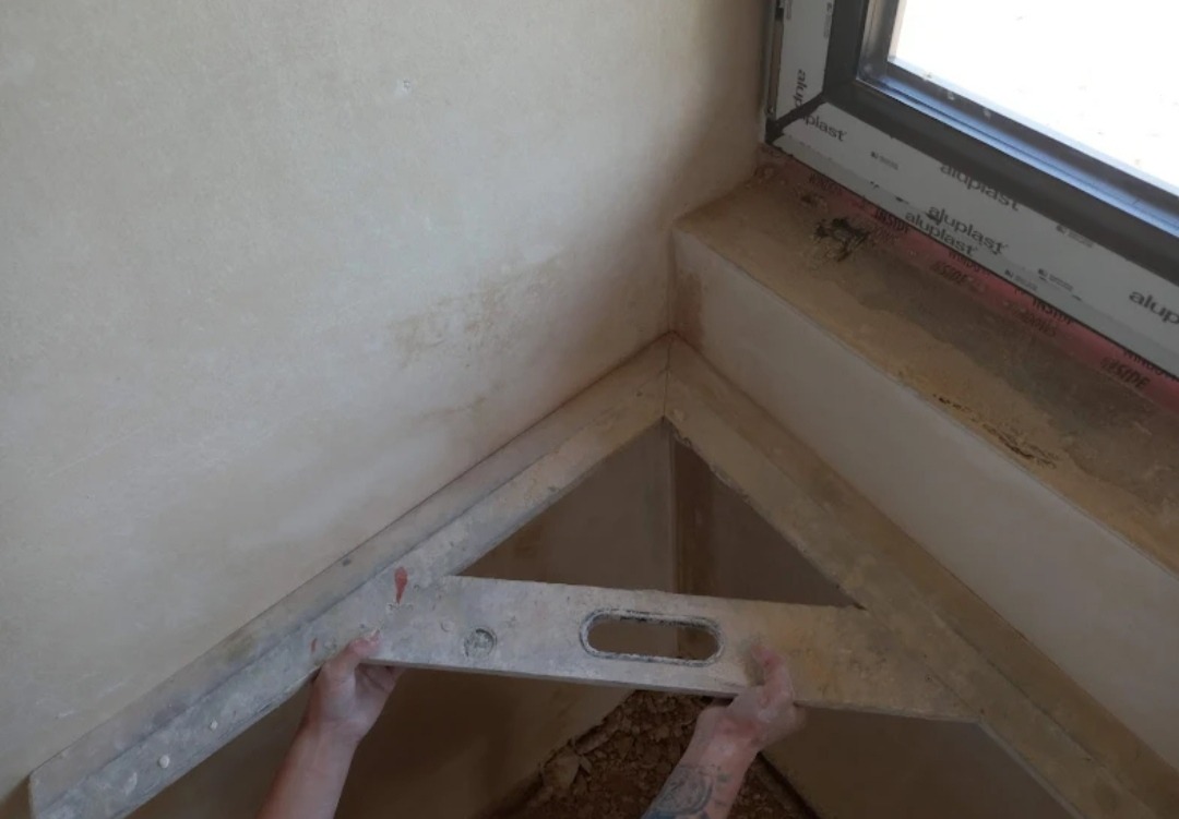 Przymiar kątowy w dłoniach, sprawdzający kąt w rogu pomieszczenia z oknem, po nałożeniu tynku maszynowego, widoczne niedoskonałości i nierówności na ścianie.