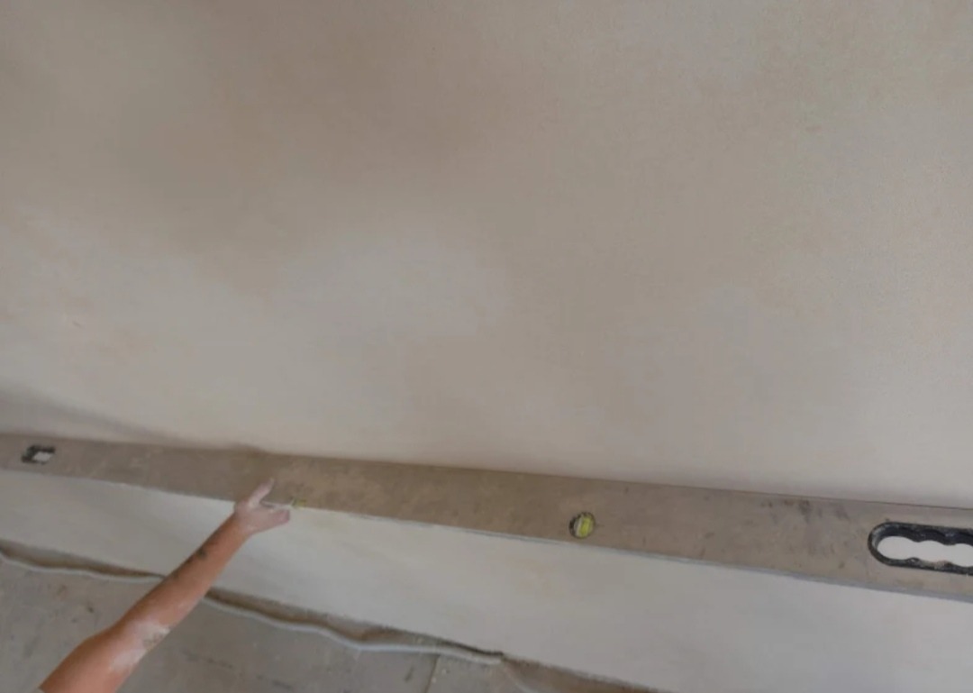 Świeżo nałożona warstwa tynku na suficie sprawdzana poziomicą przez pracownika, widoczna ręka z drobnymi zabrudzeniami tynku.