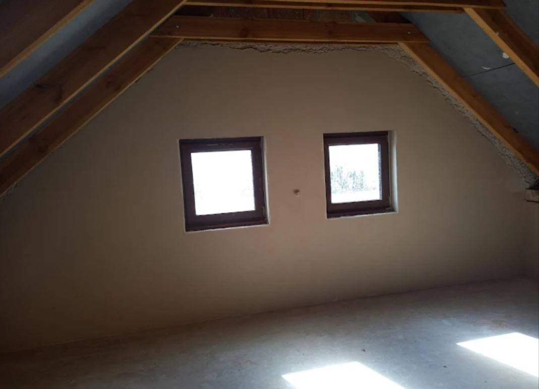 Poddasze po nałożeniu gładzi gipsowej, widoczne dwa okna w drewnianych ramach, wykończone drewniane elementy konstrukcji dachu.