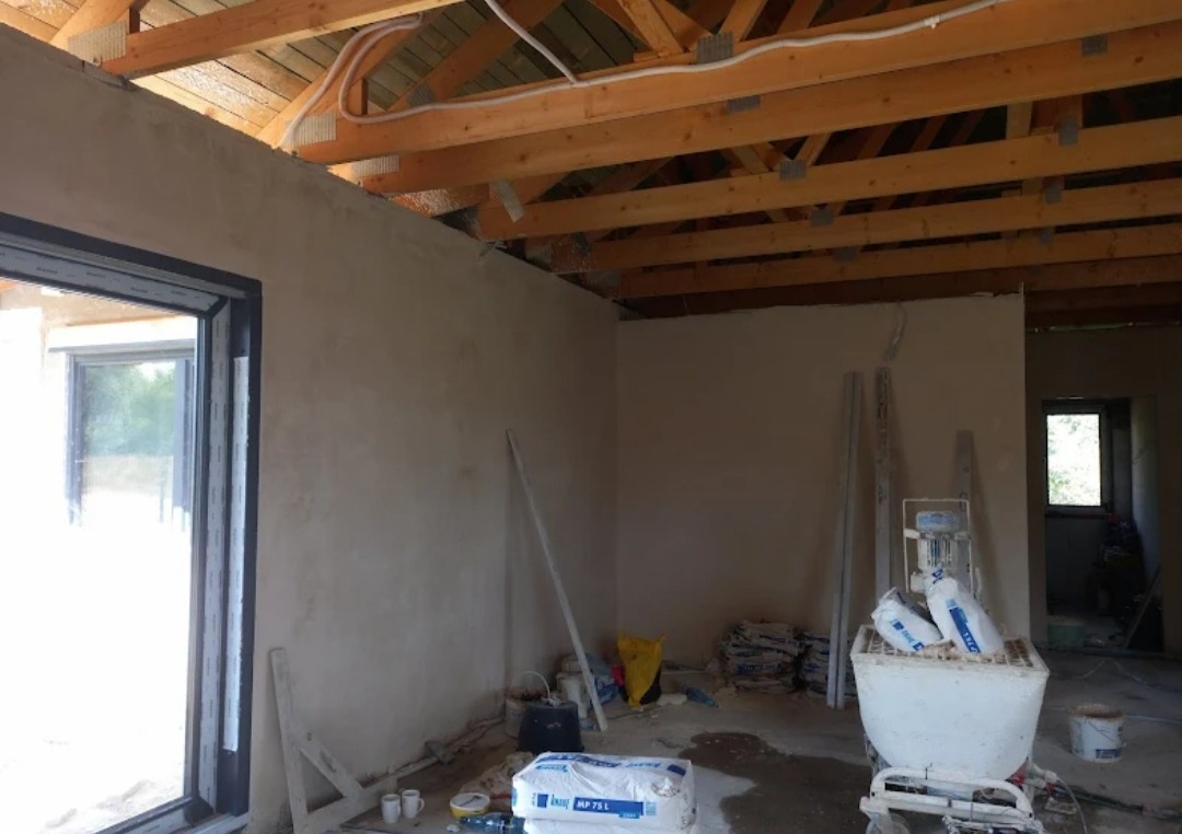 Wnętrze w trakcie prac wykończeniowych: ściany pokryte świeżym tynkiem, widoczne drewniane elementy konstrukcji dachu, worki z zaprawą oraz maszyna do nakładania tynku.