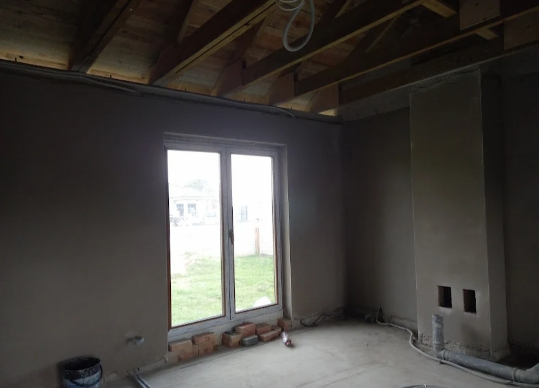 Wnętrze w stanie surowym po nałożeniu gładzi gipsowej, widoczne drewniane elementy konstrukcji dachu, okno balkonowe z widokiem na zewnątrz, u dołu ściany ułożone cegły.