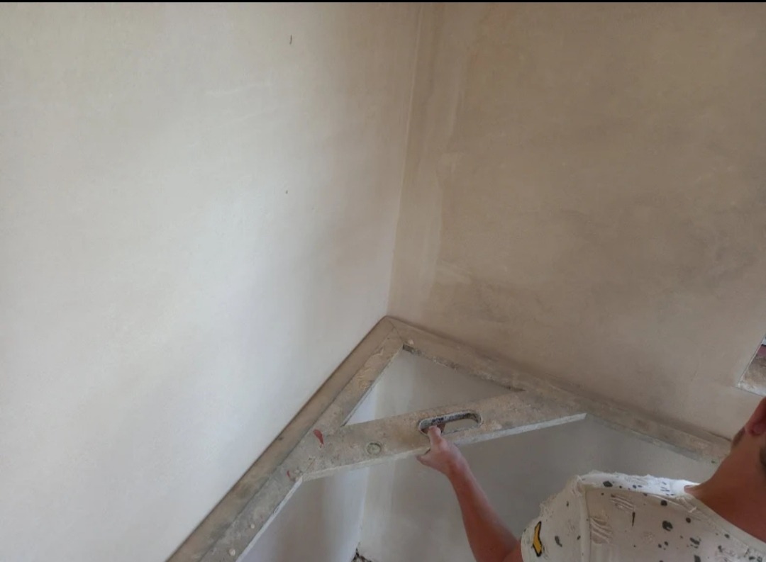 Świeżo nałożony tynk na ścianie w rogu pomieszczenia, wyrównywany łatą aluminiową przez osobę w koszulce w kropki.