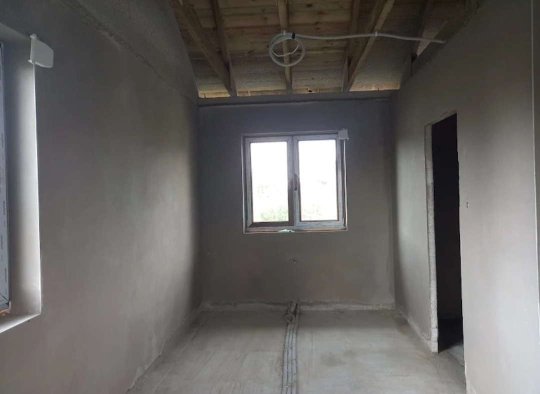 Pokój po nałożeniu tynków maszynowych: ściany i podłoga pokryte świeżym tynkiem, widoczne przewody elektryczne przy podłodze i na suficie z drewnianych desek, okno z widokiem na zewnątrz.