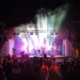 Zespół muzyczny gra koncert na oświetlonej scenie plenerowej przed zgromadzoną publicznością w nocy, z widocznymi efektami świetlnymi i delikatną mgłą.