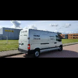 Srebrny bus transportowy Renault Master z numerem telefonu, zaparkowany na kostce brukowej przed budynkiem Netto i nowoczesnym budynkiem biurowym.