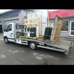 Marcel Kozłowski - Biała laweta z platformą załadunkową przewozi palety z szybami i elementami szklanymi na tle budynku stacji kontroli pojazdów.