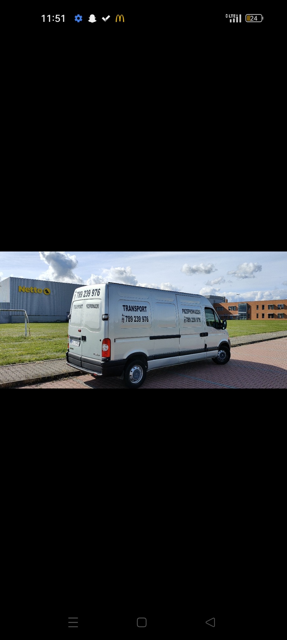 Srebrny bus transportowy Renault Master z numerem telefonu, zaparkowany na kostce brukowej przed budynkiem Netto i nowoczesnym budynkiem biurowym.