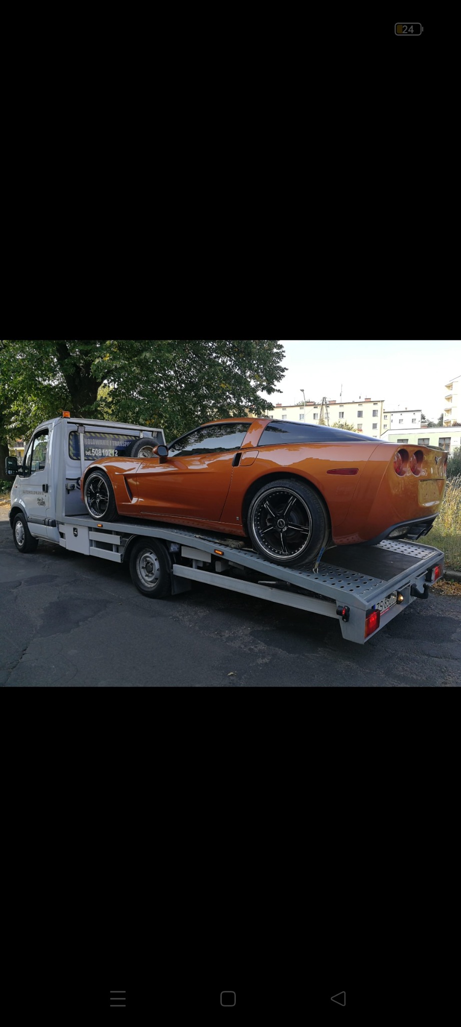 Pomarańczowy sportowy samochód Corvette na platformie lawety w trakcie transportu, widok z tyłu i boku, z numerem rejestracyjnym lawety widocznym na tablicy.