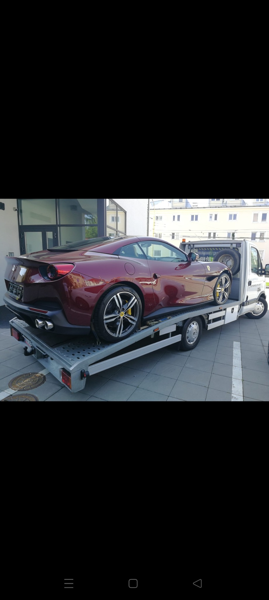 Czerwone Ferrari na platformie lawety, widok z tyłu, żółte zaciski hamulcowe, budynek w tle.