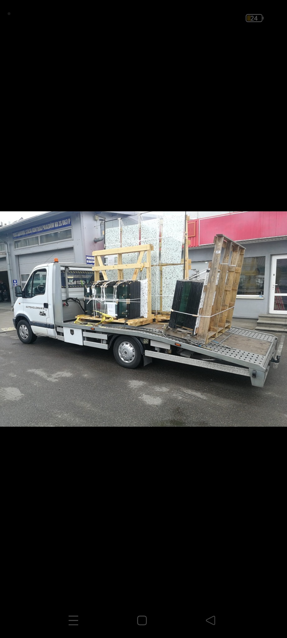 Biała laweta z platformą załadunkową przewozi palety z szybami i elementami szklanymi na tle budynku stacji kontroli pojazdów.