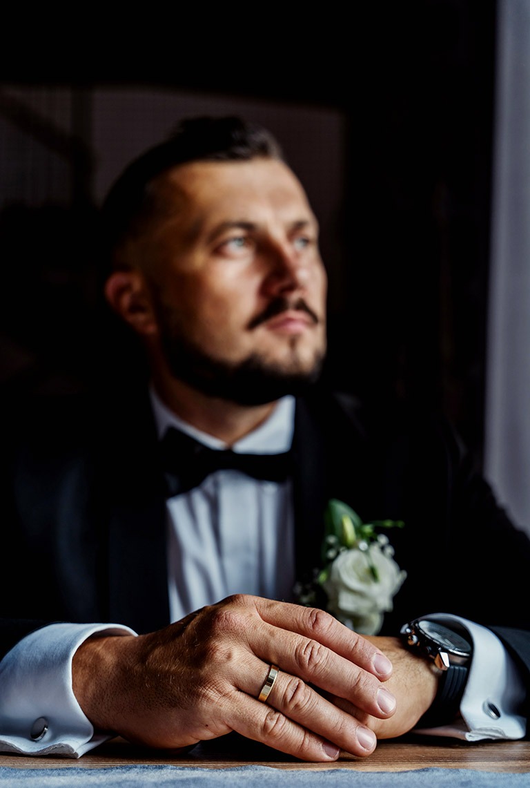 Elegancki portret mężczyzny w czarnym smokingu z muchą, spoglądającego w bok, z widocznym złotym pierścionkiem na palcu i kwiatem w butonierce.