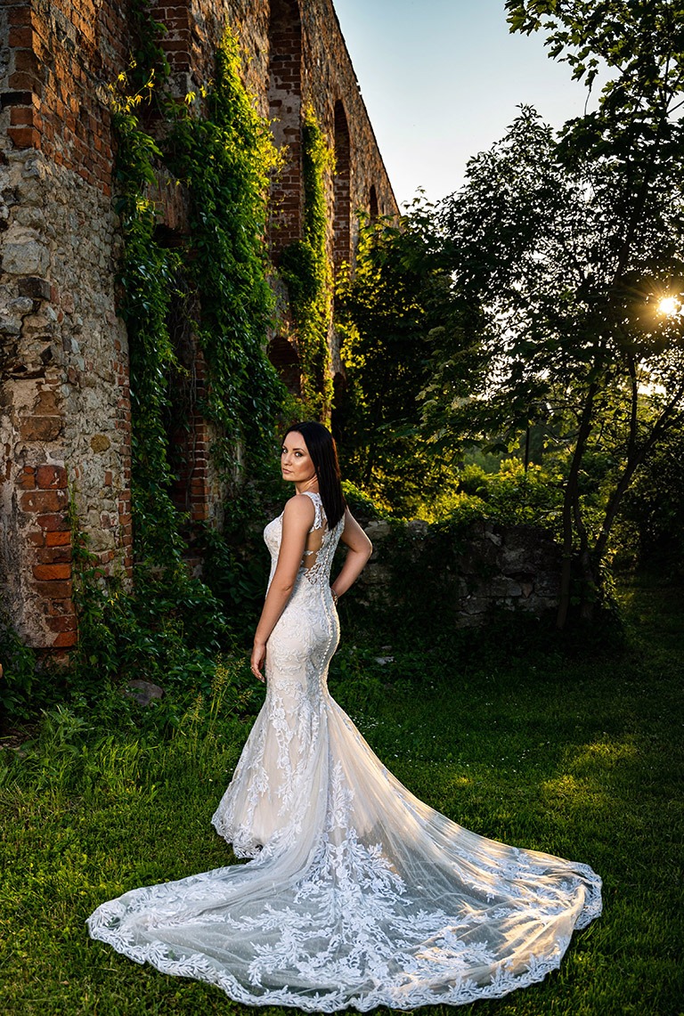 Panna młoda w koronkowej sukni z długim trenem pozuje na tle starego ceglanego muru porośniętego bluszczem w słoneczny dzień.