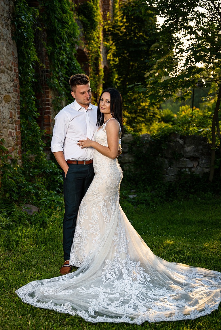 Para młoda w plenerze: panna młoda w koronkowej sukni z długim trenem, pan młody w białej koszuli i ciemnych spodniach, pozują na tle ruin porośniętych bluszczem w promieniach zachodzącego słońca.