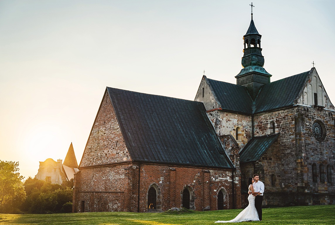 Para młoda w objęciach na tle starego, ceglanego kościoła z wieżą i krzyżem, w promieniach zachodzącego słońca.