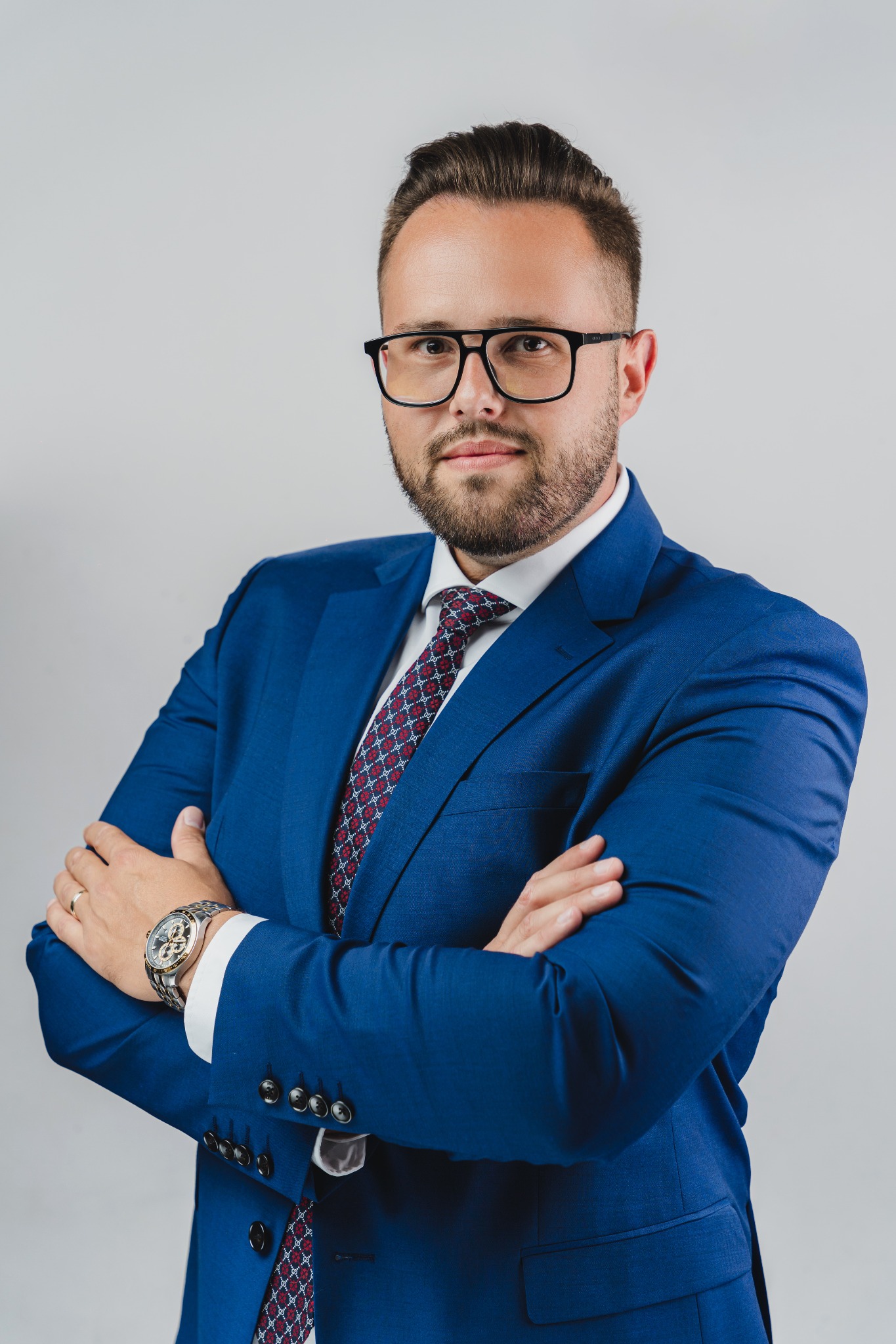 Mężczyzna w okularach i niebieskim garniturze, z zegarkiem na ręce, pozuje na jasnym tle, krzyżując ręce na piersi.
