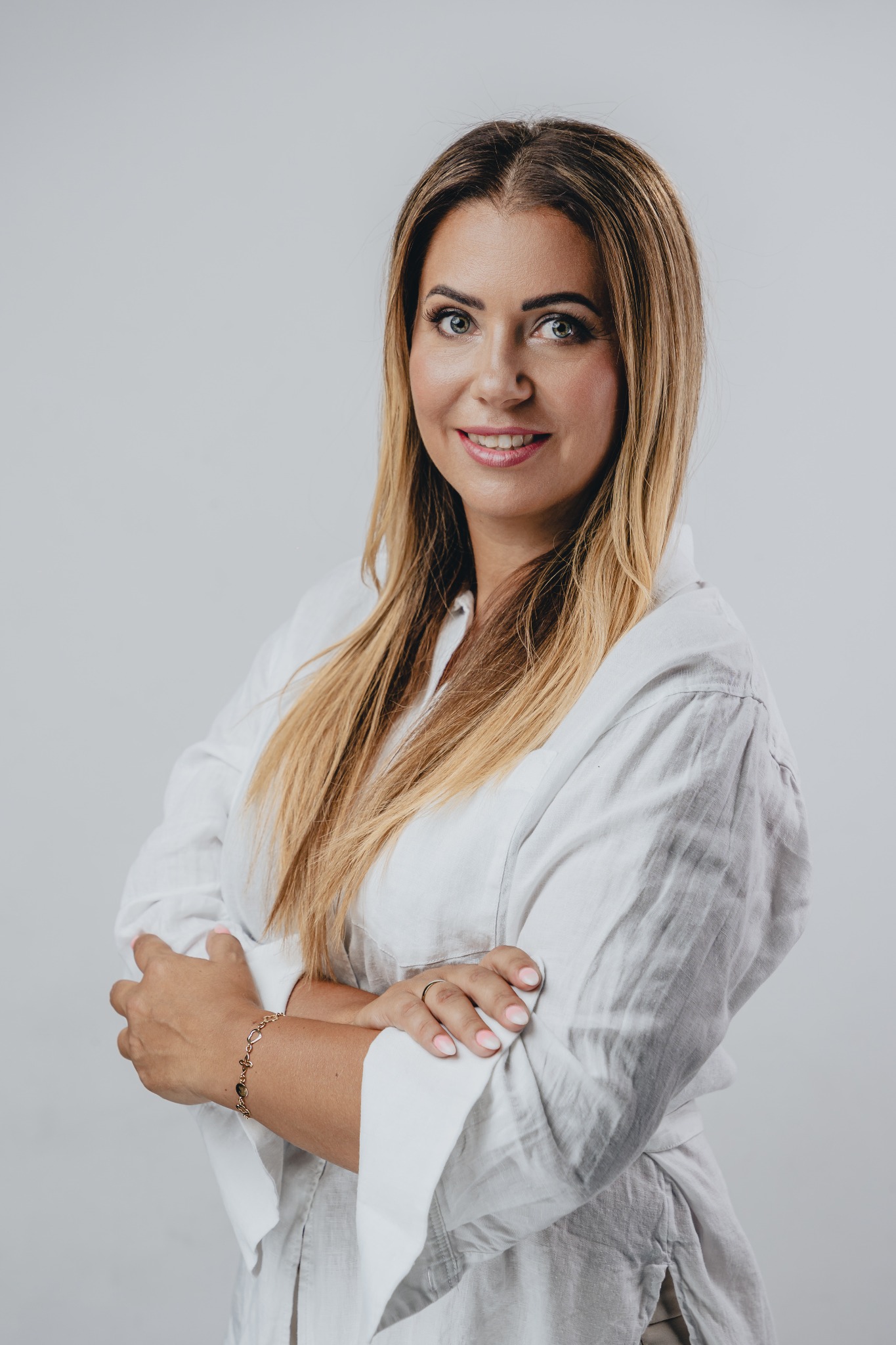 Portret uśmiechniętej kobiety w białej koszuli, ze skrzyżowanymi ramionami, na jasnym tle. Widoczny złoty pierścionek i bransoletka.