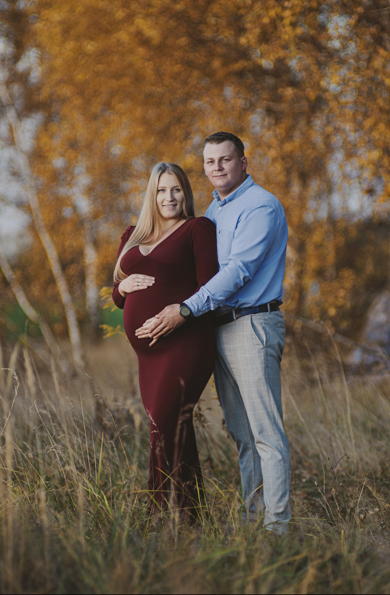 Uśmiechnięta para w jesiennej scenerii: kobieta w bordowej sukience ciążowej, mężczyzna w błękitnej koszuli i szarych spodni w kratę, oboje trzymają ręce na brzuchu.